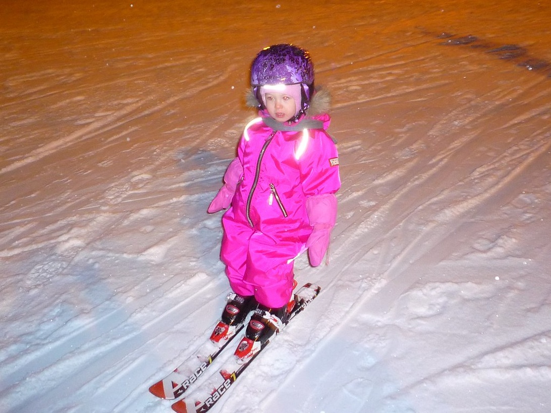 Ett litet barn i rosa skidoverall och lila hjälm står på skidor i en snöig backe på kvällen. Barnet har korrekt skidutrustning men ser ut att vara nybörjare.