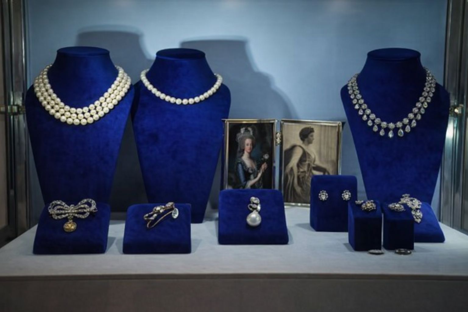 Olika smycken som halsband, broscher och örhängen visas på blåa displayer, samt två inramade porträtt.