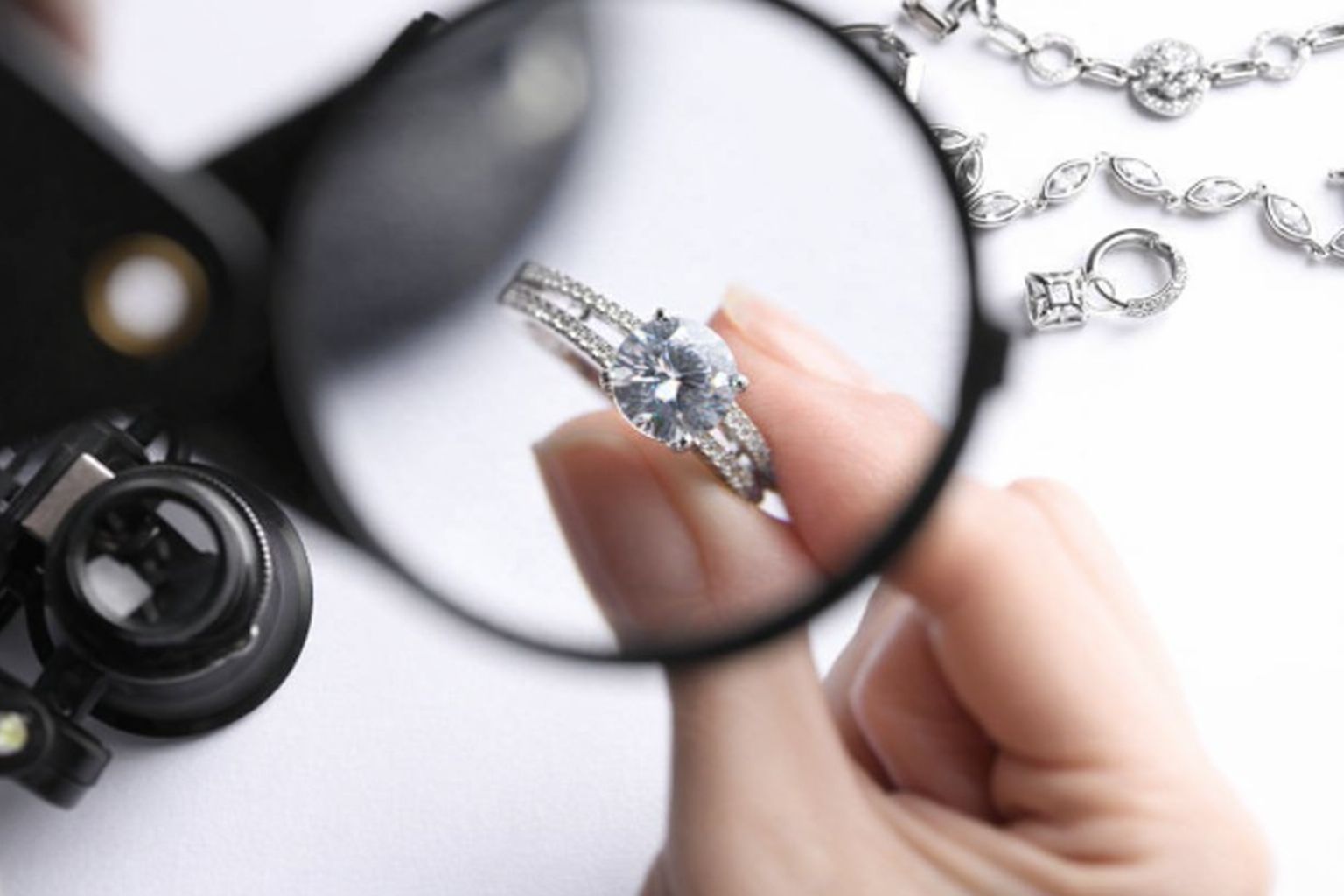 En hand håller en diamantring under ett förstoringsglas, med andra smycken i bakgrunden.